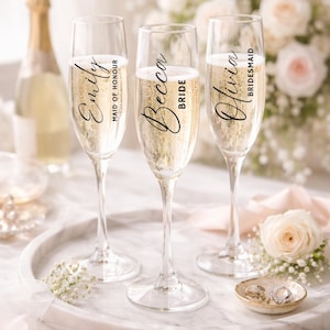 May include: Three champagne flutes filled with bubbly, each with a name and title: Emily, Maid of Honour; Becca, Bride; and Olivia, Bridesmaid. The flutes sit on a marble tray with a bottle of champagne in the background, along with flowers and a ring dish.