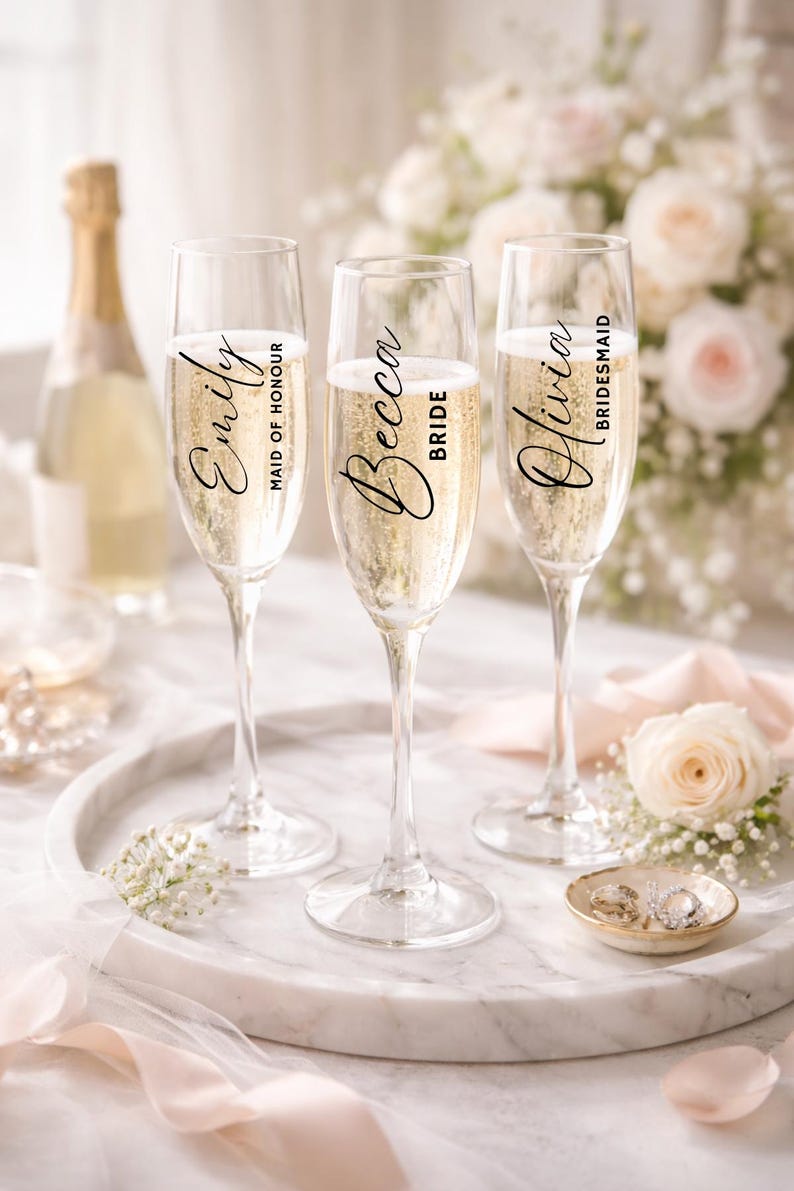 May include: Three champagne flutes filled with bubbly, each with a name and title: Emily, Maid of Honour; Becca, Bride; and Olivia, Bridesmaid. The flutes sit on a marble tray with a bottle of champagne in the background, along with flowers and a ring dish.