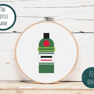 Op de afbeelding: Een kruissteekpatroon van een groene fles met een rode cirkel en een wit label. Het patroon is in een houten borduurring. De tekst "THE BOTTLE BANK" staat in een cirkel boven de borduurring en "PDF ONLY" staat in een cirkel onder de borduurring.