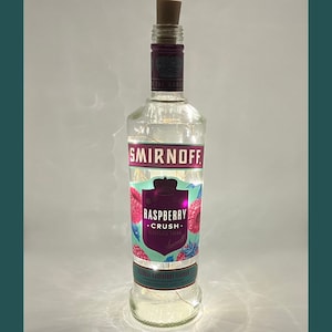 May include: A clear glass bottle with a cork top and a label that says "Smirnoff Raspberry Crush" with a pink and green design. The bottle is decorated with white fairy lights.