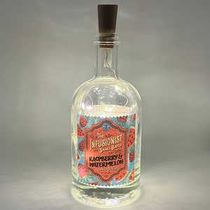 May include: A clear glass bottle with a brown cork stopper, filled with string lights. The label reads "The Infusionist Small Batch Raspberry & Watermelon Gin Liqueur". The bottle is illuminated from within, creating a warm glow.