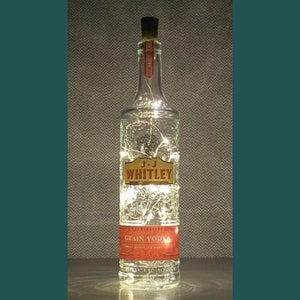 May include: A clear glass bottle with a cork top and a label that reads "J.J. Whitley Grain Vodka". The bottle is filled with white fairy lights.