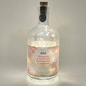 May include: A clear glass bottle with a cork stopper and a label that reads "Rhubarb & Scottish Raspberry Flavour Gin". The bottle is decorated with a pink and white floral design and contains a string of white lights.