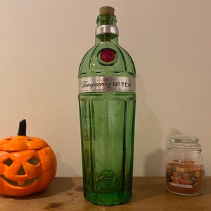 Pu&ograve; includere: Una bottiglia di vetro verde con un tappo di sughero e un'etichetta che recita "Tanqueray No. Ten" &egrave; appoggiata su una superficie di legno. Sulla superficie si trovano anche una piccola zucca arancione e un barattolo di vetro con un coperchio e un'etichetta che recita "Woodwick".