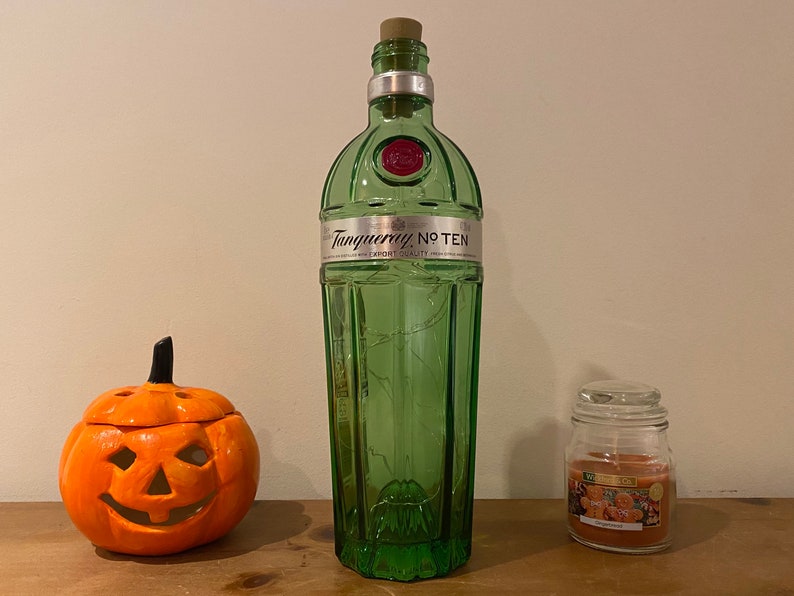 Pu&ograve; includere: Una bottiglia di vetro verde con un tappo di sughero e un'etichetta che recita "Tanqueray No. Ten" &egrave; appoggiata su una superficie di legno. Sulla superficie si trovano anche una piccola zucca arancione e un barattolo di vetro con un coperchio e un'etichetta che recita "Woodwick".