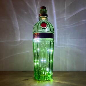 Pu&ograve; includere: Una bottiglia di vetro verde con un tappo di sughero e luci bianche fatate all'interno. La bottiglia ha un'etichetta che dice "Tanqueray London Dry Gin".