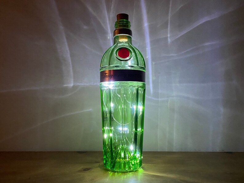 Pu&ograve; includere: Una bottiglia di vetro verde con un tappo di sughero e luci bianche fatate all'interno. La bottiglia ha un'etichetta che dice "Tanqueray London Dry Gin".