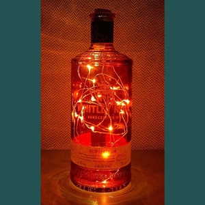 May include: A clear glass bottle with a label that reads "Whitley Neill Handcrafted Gin" and "Raspberry Gin". The bottle is filled with warm white fairy lights.