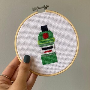 May include: A green and black cross-stitch embroidery of a liquor bottle with a red circle and white label. The embroidery is framed in a wooden hoop.