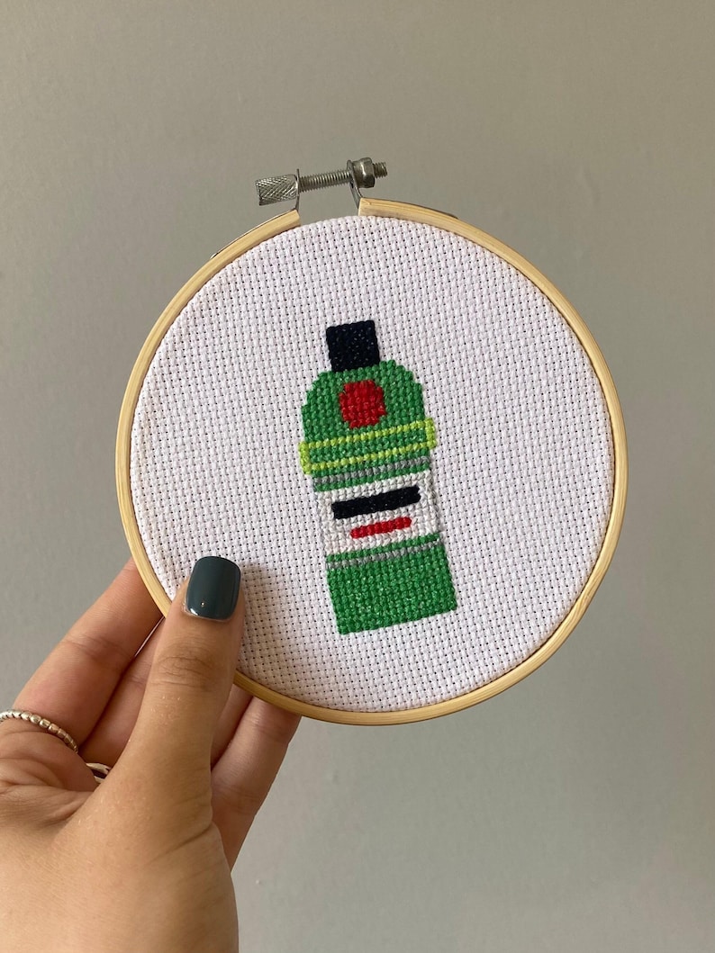 May include: A green and black cross-stitch embroidery of a liquor bottle with a red circle and white label. The embroidery is framed in a wooden hoop.