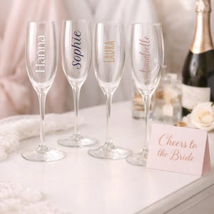 May include: Four champagne flutes with names Hanna, Sophie, Laura, and Annabelle printed on them. A small card reads "Cheers to the Bride." A bottle of champagne and a white rose are in the background. The flutes are on a white surface.