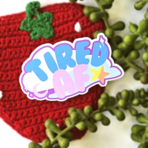 Puede incluir: Una pegatina con las palabras "TIRED AF" en azul, rosa y amarillo, con un contorno blanco. La pegatina está colocada sobre una fresa roja de ganchillo con detalles blancos y hojas verdes. Plantas suculentas verdes en el fondo.