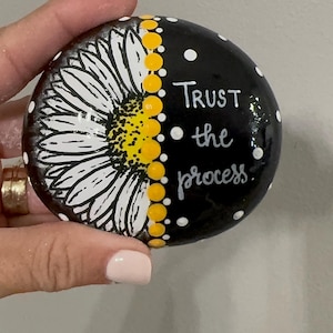May include: Hand-painted, round stone featuring a sunflower design. Half of the stone displays a white and yellow sunflower, while the other half is black with white polka dots and the words "Trust the process."