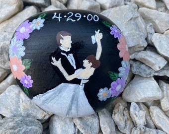 May include: A hand-painted black stone featuring a couple dancing, surrounded by floral accents in pink and purple. The date "4.29.00" is written in white. The stone is resting on a bed of gray rocks.