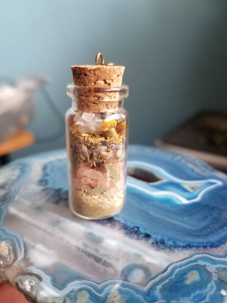 Może przedstawiać: Mała szklana butelka z korkowym korkiem wypełniona suszonymi ziołami, kryształami i złotym metalowym wisiorkiem. Butelka stoi na niebiesko-białym agacie.