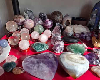 Op de afbeelding: Een verzameling gepolijste kristallen bollen, hartvormige stenen en ruwe mineraalclusters in diverse kleuren, waaronder paars, roze, groen en transparant. Het assortiment bevat gladde, bolvormige stenen op kleine standaards en rotsformaties.