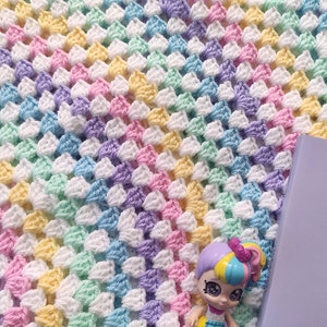 May include: A rainbow crochet blanket with a pastel colour scheme. The blanket is made with a granny square stitch and features shades of pink, yellow, blue, green, and purple.