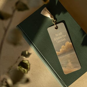 Peut inclure: Un marque-page avec un pompon repose sur un livre fermé, vert foncé. Le marque-page présente le texte "Be still & know that I am God" au-dessus d'une image de paysage de nuages et d'eau. Le pompon est beige clair.