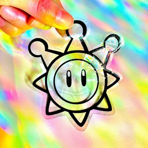 May include: A holographic sticker of a cartoon sun with a face and a crown. The sticker is black and white with a rainbow iridescent finish.