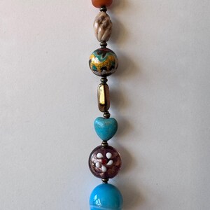 Puede incluir: Una hebra de cuentas de colores con una variedad de texturas y patrones. Las cuentas incluyen una marrón, una blanca, una azul y dorada, un corazón turquesa, una morada y blanca y una azul y blanca.