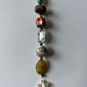 以下が含まれることがあります： 緑と青のガラスビーズ、銀色の金属ビーズ、赤と緑のガラスビーズ、透明なガラスビーズ、白とピンクの磁器ビーズ、茶色の瑪瑙ビーズ、白と虹色の貝殻ビーズなど、さまざまな色と質感のビーズでできたチャームです。