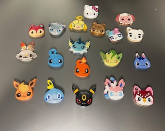 Op de afbeelding: Een verzameling van 18 kleurrijke karaktercharms, met populaire cartoon- en videogamepersonages. De charms zijn gemaakt van plastic en hebben een verscheidenheid aan ontwerpen, waaronder een roze Hello Kitty, een blauw-wit Pokémon-personage en een groen-geel personage uit de Animal Crossing-videogame.