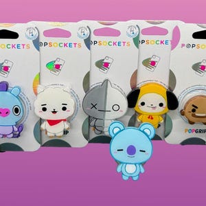 Puede incluir: Una colección de PopSockets con diseños de personajes de dibujos animados. Los PopSockets están adheridos a tarjetas blancas con el logotipo de PopSockets y un agujero negro para colgar. Los personajes incluyen un conejo rosa, un oso azul y otros.