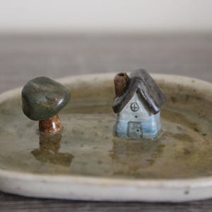 May include: A ceramic miniature landscape scene featuring a small blue house with a dark roof and a tree on a round, shallow dish. The dish has a light green and beige color scheme.