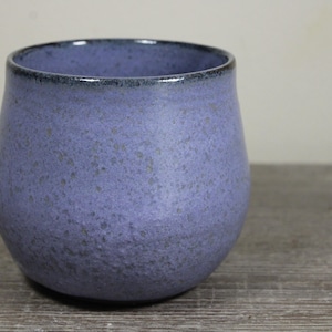 May include: A rounded ceramic mug with a speckled lavender and purple glaze. The exterior features a textured, mottled finish with a dark, smooth rim. The vessel has a wide, bulbous body that tapers slightly toward the top opening.
