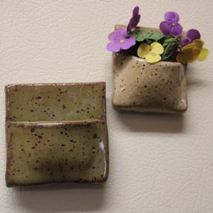 Op de afbeelding: Twee keramische wandplantenbakken. Eén is leeg, de andere bevat paarse en gele bloemen. De plantenbakken zijn rechthoekig met een gespikkelde, aardse afwerking. Ze zijn gemonteerd op een crèmekleurig oppervlak.