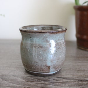 Może przedstawiać: Mały, ręcznie robiony ceramiczny garnek o zaokrąglonym kształcie. Garnek ma nakrapianą, ziemistą paletę kolorów brązu, szarości i bladoniebiesko-zielonego. Obręcz jest ciemniejsza brązowa, a podstawa ciemniejsza brązowa. Garnek jest prawdopodobnie przeznaczony do roślin.