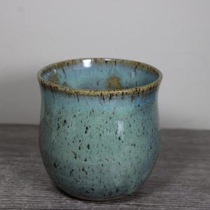 May include: A ceramic mug with a rounded body and a wide opening. The exterior features a speckled teal and blue glaze with dark brown flecks throughout. The rim is finished with a smooth, dark brown band that transitions into the blue glaze.