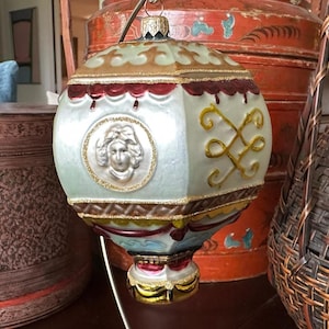 May include: A decorative Christmas ornament shaped like a hot air balloon. The ornament is light blue with gold and red accents, featuring a woman's face in a circular frame. The top and bottom have gold glitter trim.