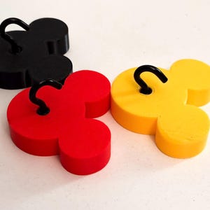 May include: Three Mickey Mouse-shaped hooks in black, red, and yellow. Each hook has a curved black metal hook attached to the top. The hooks are arranged on a white surface.