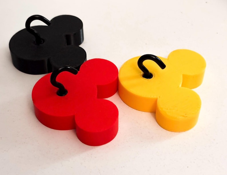 May include: Three Mickey Mouse-shaped hooks in black, red, and yellow. Each hook has a curved black metal hook attached to the top. The hooks are arranged on a white surface.
