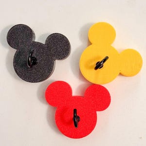 May include: Three Mickey Mouse-shaped wall hooks in black, yellow, and red. Each hook has a black metal loop for hanging. The hooks are made of wood and are arranged on a white surface.
