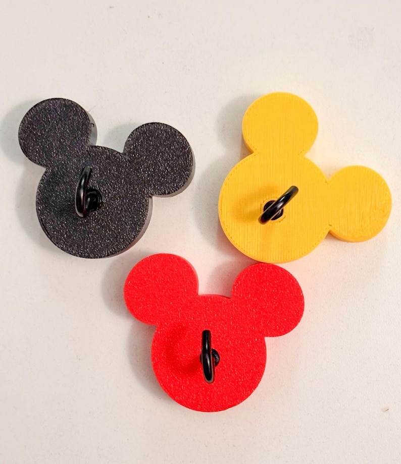 May include: Three Mickey Mouse-shaped wall hooks in black, yellow, and red. Each hook has a black metal loop for hanging. The hooks are made of wood and are arranged on a white surface.