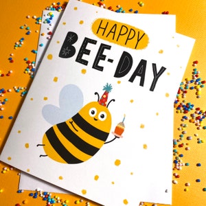 May include: A white card with a bee illustration and the text "Happy Bee-Day". The bee is yellow and black with a party hat and is holding a cupcake.