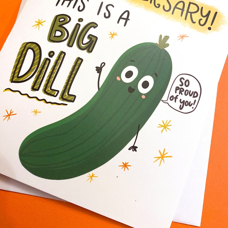 Pu&ograve; includere: Un biglietto bianco con un cetriolo di cartone animato verde con un viso e delle braccia. Il cetriolo dice "So proud of you!" in una nuvola di dialogo. Il biglietto dice anche "This is a big dill!" in lettere gialle e nere.