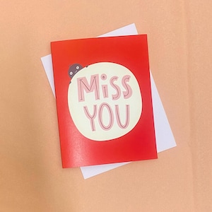 Może przedstawiać: Czerwona kartka z życzeniami z różowymi słowami "Miss You" w białym okręgu. Mała postać z kreskówki wygląda z lewego górnego rogu. Biała koperta znajduje się za kartką.