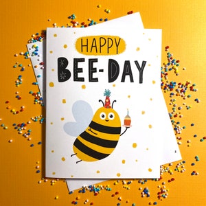 May include: A white card with a yellow and black bee wearing a party hat and holding a cupcake. The card says "Happy Bee-Day" in black letters.