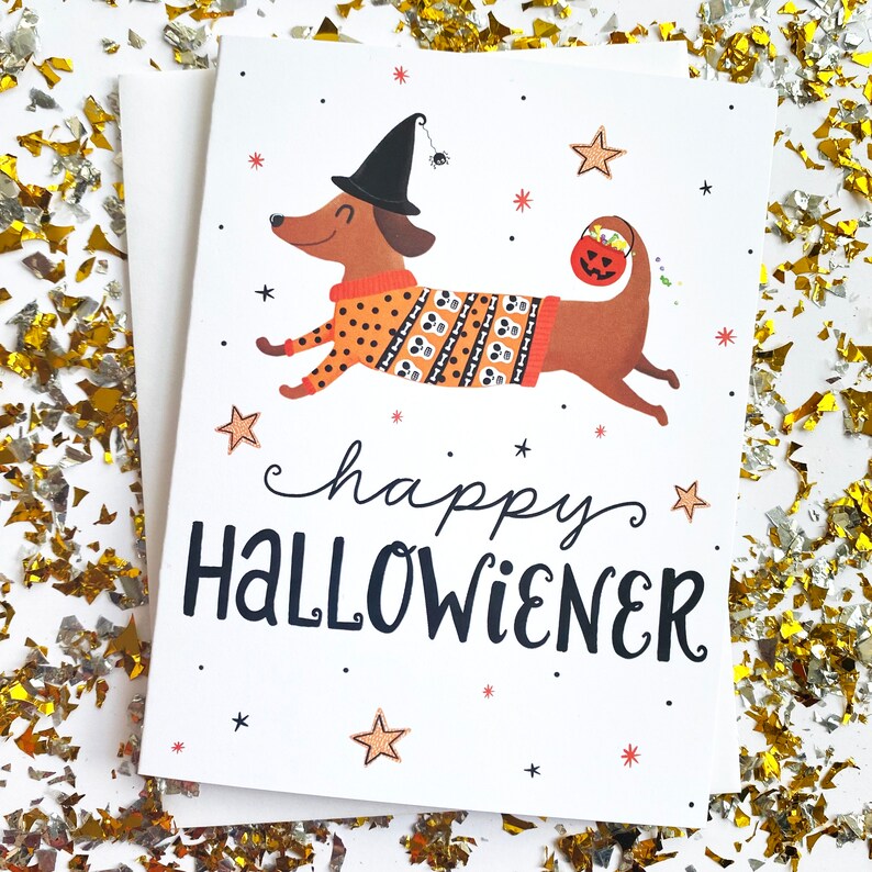 Op de afbeelding: Een witte kaart met een cartoon teckel die een heksenhoed en een trui met schedels draagt. De teckel houdt een pompoen-emmer vast. De tekst op de kaart luidt "happy HALLOWIENER".