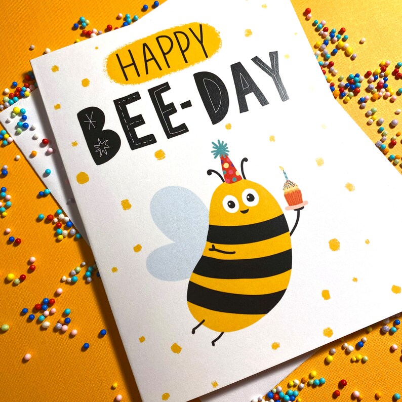 May include: A white card with the text "Happy Bee-Day" in black and a cartoon bee with a party hat holding a cupcake. The bee is yellow and black with a white face and black eyes.