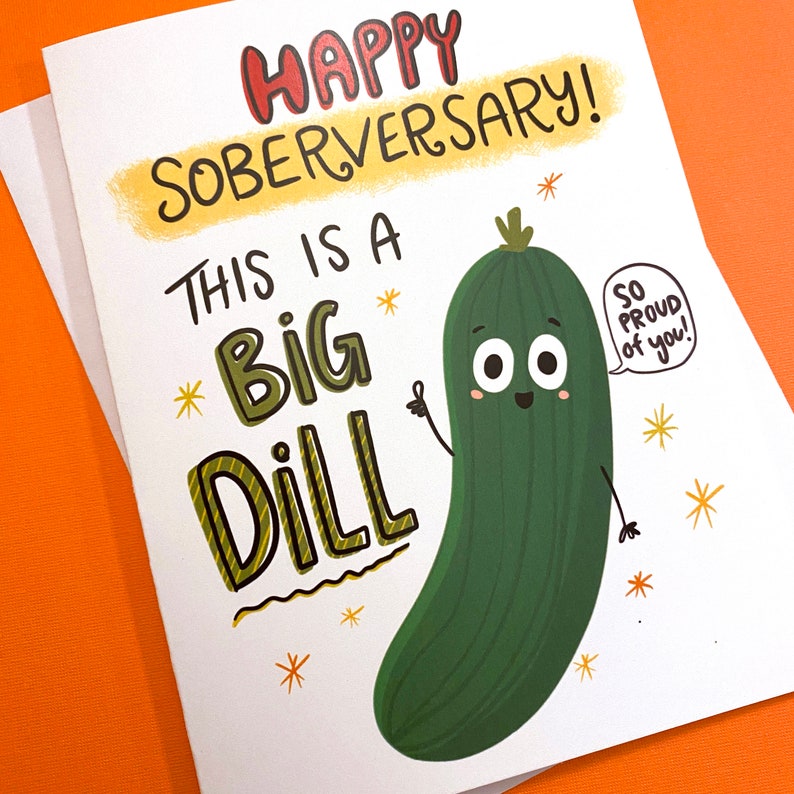 Pu&ograve; includere: Un biglietto bianco con un cetriolo di cartone animato verde su di esso. Il biglietto dice "Happy Soberversary! This is a Big Dill" e "So Proud of You!" in un fumetto sopra il cetriolo.