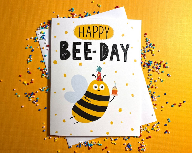 May include: A white card with a yellow and black bee wearing a party hat and holding a cupcake. The card says "Happy Bee-Day" in black letters with white polka dots.