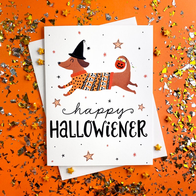 Op de afbeelding: Een witte kaart met een schattig cartoon teckel die een heksenhoed en een trui met schedelpatroon draagt. De teckel houdt een emmer snoep vast. De kaart zegt "happy HALLOWIENER" in zwarte schrift.