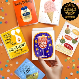 May include: A selection of humorous birthday cards with various illustrations and text. The cards feature images of a mayonnaise jar, ice cream cone, egg, cocktail, and fish. The cards are made in the USA.