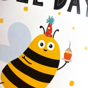 May include: A yellow and black bee wearing a colorful party hat holds a cupcake with a single candle. The bee has big, round eyes and a happy expression. The background is white with gold glitter.