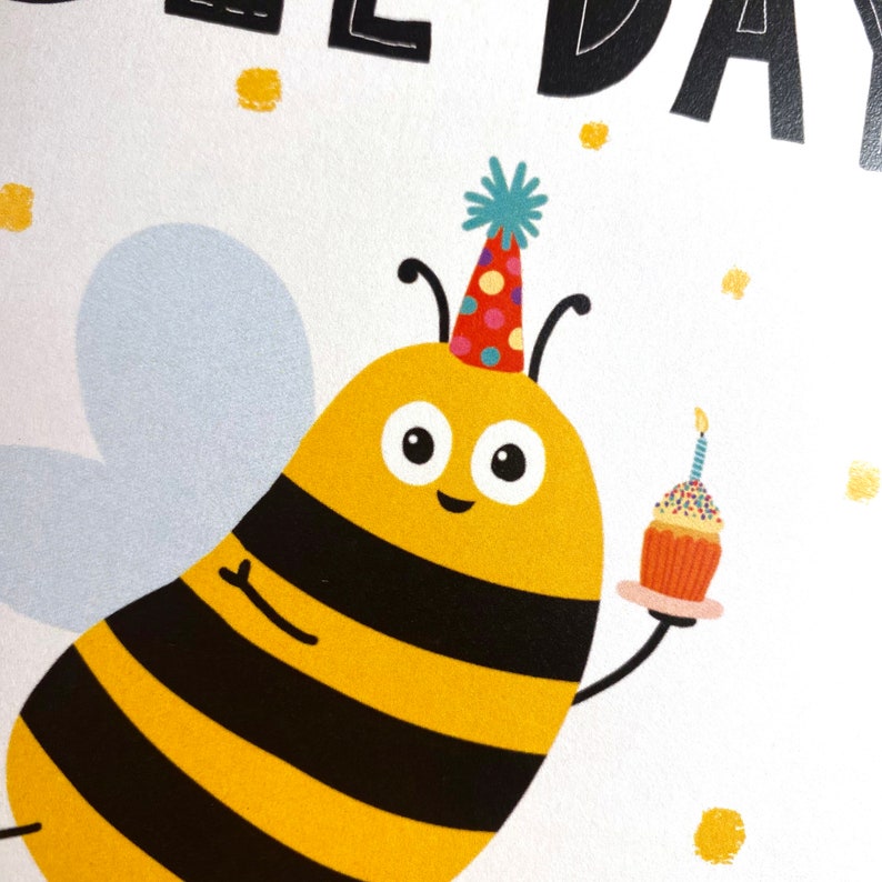 May include: A yellow and black bee wearing a colorful party hat holds a cupcake with a single candle. The bee has big, round eyes and a happy expression. The background is white with gold glitter.