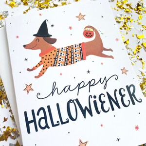 Op de afbeelding: Een witte kaart met een bruine teckel die een heksenhoed en een trui met een schedel- en botpatroon draagt. De teckel houdt een pompoen-emmer vast en de tekst "happy hallowiener" is in zwart geschreven.